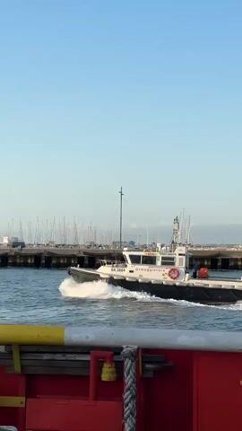 A white and black boat with the words "ARMeggiater" on its side is moving through the water, creating a wake. The boat has a white cabin and a red life preserver is visible on its side. Behind the boat, a marina with many sailboats docked is seen, along with a clear blue sky. In the foreground, a red structure with yellow and white railings is partially visible. There are at least two people on the boat. The water appears choppy.
