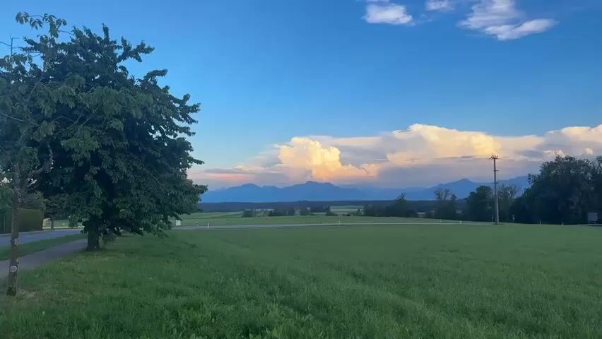 This is a landscape video with a wide shot of a green meadow under a blue sky with white clouds. In the distance, there are mountains and large cumulus clouds that are lit by the setting sun, creating a warm orange and pink glow. A few trees are visible on the right side of the frame, and a single tree stands on the left. A road and a utility pole are also present in the scene. The overall atmosphere is peaceful and serene.