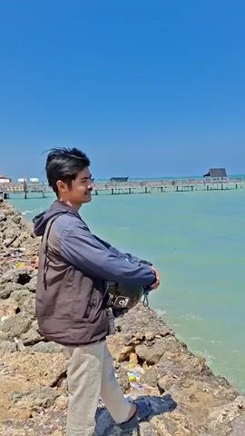 A young man with dark hair and a mustache stands on a rocky shore, looking out at a blue sky and turquoise water. He is wearing a dark vest over a blue hoodie and light-colored pants. He holds a black helmet in his arms. In the background, a wooden pier extends into the water, with structures on it and in the distance. The shore is covered in rocks and some debris. The sun is shining brightly, casting a shadow of the man onto the rocks.