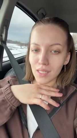 A young woman is sitting in the driver's seat of a car. She is wearing a brown zip-up hoodie over a white t-shirt, and her seatbelt is fastened. She has light-colored eyes and shoulder-length light brown hair, partially pulled back. She is wearing a simple gold ring and rectangular gold earrings. The woman is looking directly at the camera, talking and gesturing with her hand. The background shows the car's interior and a blurred view of a landscape outside the window. There appears to be some snow on the ground. The car radio is playing music.