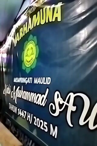 The video shows a speaker at a Maulid Nabi Muhammad SAW celebration in 2025. The speaker, dressed in traditional Muslim attire, stands at a wooden podium. Seated in rows of plastic chairs are children, most of whom are wearing traditional Muslim attire. A banner in the background reads "Memperingati Maulid Nabi Muhammad SAW, Tahun 1447 H/2025 M." The event is held under a white tarp, decorated with festive lights. The atmosphere is lively and celebratory.