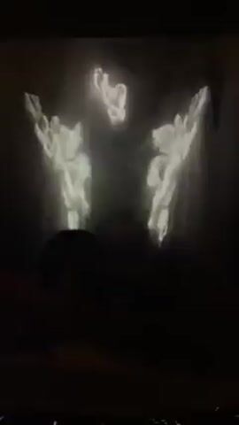 The video shows a dark room with three bright, ghostly figures projected on the wall. They appear to be of people with their arms raised. A person is visible in silhouette in the foreground, seated in front of the projection.