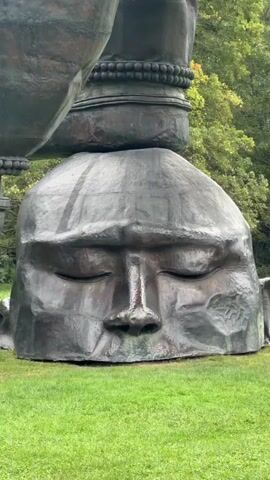 A large, dark-colored sculpture of a stylized human head with closed eyes is resting on green grass. On top of the head is another part of the sculpture, a large, dark-colored leg with a band around the ankle, which appears to be in motion. The leg is elevated, and a foot is resting on a wooden support. The background consists of trees with green and yellow foliage, suggesting an outdoor setting, possibly a park or sculpture garden. The sky is overcast.
