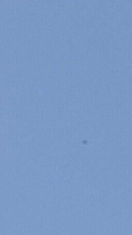 The video shows a small, dark object in a blue sky, initially centered in the frame. The camera angle then shifts to show a dark, possibly dirty, car roof in the foreground with the clear, blue sky above. The object in the sky is still visible, but less prominent.
