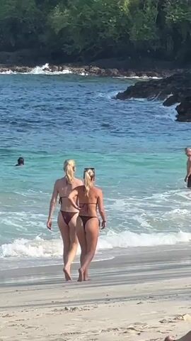 Two women in bikinis walk along a sandy beach towards the ocean. Waves lap at the shore. In the water, several people are visible. To the left, a person in the water is swimming. Further out, a man stands in the water. To the right of the women, a child in a green bathing suit is standing in the water. The background consists of a rocky coastline with lush green foliage.
