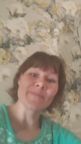 A woman with short brown hair and bangs smiles and makes faces at the camera. She is wearing a teal shirt with a white and grey floral pattern. The background is a floral wallpaper in shades of cream, yellow, and green.