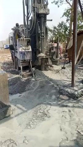 The video shows a drilling rig in operation, with a person visible on the platform of the truck-mounted machine. The ground surrounding the rig is covered in a slurry of mud and rock fragments, indicating active drilling. There is a concrete structure and a makeshift shelter in the background, along with trees and a clear sky suggesting an outdoor work environment. The rig's mechanical components are in motion, and the sound of the drilling operation is audible.