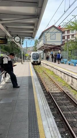 A train pulls into a station.  A man in a black jacket and jeans stands on the platform looking at his cell phone. There is a clock on the station wall behind him, and a trash can next to the platform edge.  Across the tracks, another platform has people waiting and blue benches. The train is white and silver with blue trim.  The train's designation, 913, is visible on the front. The train says "Bilbade" on the side. The building across the tracks is light blue and white.