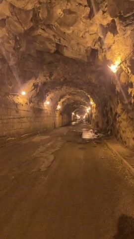 The camera slowly advances through a dimly lit tunnel. The tunnel walls and ceiling are composed of rough, hewn rock, with occasional sections of what appears to be man-made masonry. Small, warm-toned lights are spaced along the walls, providing minimal illumination. The road surface is asphalt but is cracked and shows evidence of standing water. A few indistinct figures and vehicles are visible at the far end of the tunnel, their lights creating a point of brightness in the distance.