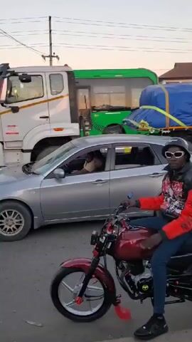 The video shows a street scene with various vehicles and pedestrians. A man on a red motorcycle with custom accessories is in the foreground. He is wearing a black helmet, sunglasses, a jacket with red sleeves, blue jeans, and black shoes. Around him are other motorcycles and cars, including a silver Subaru and a green truck with a blue tarp-covered load. The traffic light is green, allowing vehicles to proceed. Pedestrians, including a person wearing a white shirt with a backpack, are also visible on the side of the road. In the background are buildings and trees, suggesting an urban environment. The sky appears to be getting dark, indicating it might be late afternoon or early evening.