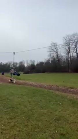 A man is riding a zipline outside in a grassy field. He wears a grey jacket, jeans and black shoes. A dog with black, tan and white markings runs below him barking. There are trees and a grey sky in the background.