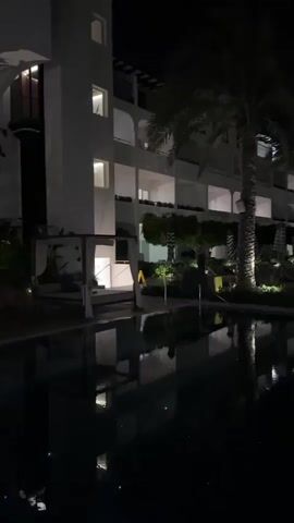 This media file depicts a nighttime scene at a resort or hotel complex in Playa Del Sol-Villicana, Spain. The main subject is a tranquil courtyard or pathway illuminated by artificial lights, bordered by a reflecting pool and a multi-story building.

The building, a white structure with multiple balconies and windows, appears to be a hotel or resort. Lights are visible in some of the windows, suggesting occupancy. Palm trees are a prominent feature of the landscape, with their fronds silhouetted against the night sky and illuminated by spotlights. The reflecting pool in the foreground mirrors the building and the palm trees, creating a serene and slightly mystical atmosphere. Lounge chairs and umbrellas are visible near the pool, indicating a recreational area.

The time of day is clearly night, evidenced by the darkness of the sky and the reliance on artificial lighting. The weather appears calm, with the still water of the pool providing clear reflections. The overall mood is peaceful and inviting, suggesting a relaxing evening at a holiday destination.

There is no visible text in the media file. The scene evokes a sense of tranquility and luxury, typical of a resort environment.