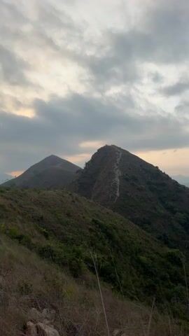 The video presents a scenic panoramic view of a mountainous landscape, characteristic of the Bac Natural Heritage Area in Trang, Vietnam. The scene opens with a vista of two prominent hills, the nearer one featuring a distinct, winding path along its ridge. The sky above is partly cloudy, casting a soft, warm light that suggests either early morning or late afternoon. The immediate foreground is covered in dry grass and scattered rocks.

As the camera slowly pans from left to right, it reveals an extensive panorama of rolling hills and mountains, densely covered in lush green vegetation and trees. The lighting subtly brightens during the pan, transitioning the sky from a gentle, overcast appearance to a clearer, light blue hue. Further into the distance, successive layers of hills fade into a soft haze, culminating in what appears to be a body of water in the far distance on the right, possibly the sea.

The pan concludes by showcasing a valley below, where signs of human activity, such as cultivated areas or small settlements and roads, are visible amidst the natural terrain. The overall ambiance is one of serene natural beauty and expansive vistas. No people or visible text are present in the footage.