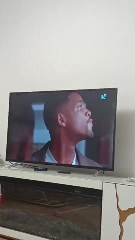 A wide shot shows a flat-screen television mounted on a white media console in a room with white walls. The television displays a movie scene featuring Will Smith in a close-up shot on the left side of the screen. On the right side of the screen, a logo with the letter "K" inside a circle is visible. The media console has a dark glass top with a patterned design and a black electronic device below the television screen. To the left of the television, a plastic bottle with a red label is partially visible on the console. The scene transitions to a woman holding a small animal. The room appears to be dimly lit.