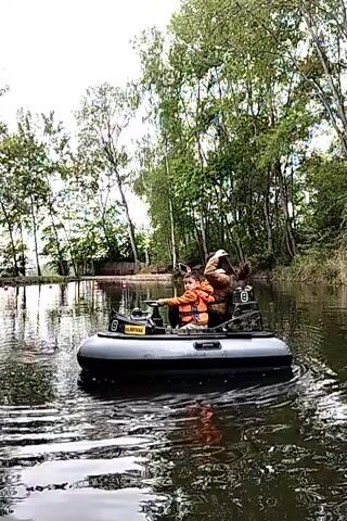 A woman and a young child are riding in a small gray inflatable boat on a lake. The child is wearing a bright orange life vest and is sitting in the front of the boat, holding the steering controls. The woman sits behind the child, holding up a cell phone as if taking a picture. The boat has the number "8" on its side and the word "FAIRPARK" printed on a yellow plaque. The lake is surrounded by green trees and reflected in the water. The sky is overcast. The boat is moving slowly across the calm water.