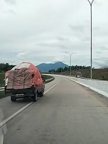 A vehicle, possibly a car, is driving on a highway behind a small truck. The truck is heavily loaded and covered with a tarp secured with ropes. A mountain is visible in the background. The sky is overcast. The highway is mostly empty with visible lane markings and guardrails. There are streetlights along the side of the road. There are some trees in the distance. The video appears to be shot from inside the trailing vehicle.