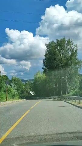 The video shows a view from a moving vehicle driving on a paved road. The road has a single yellow dashed line painted down the center. The road curves to the right. On either side of the road, there are trees and green foliage. In the distance, there appears to be a small building. The sky is blue with large white clouds. Overhead, there are power lines.