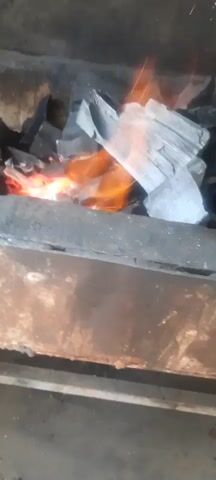 A close-up, top-down view of charcoal burning in a fire pit. The charcoal pieces are dark gray and appear to be breaking apart. Orange flames lick at the charcoal, and a few sparks fly upwards. The fire pit itself is made of dark metal, with some rust visible. The overall environment is dark and appears to be outdoors.