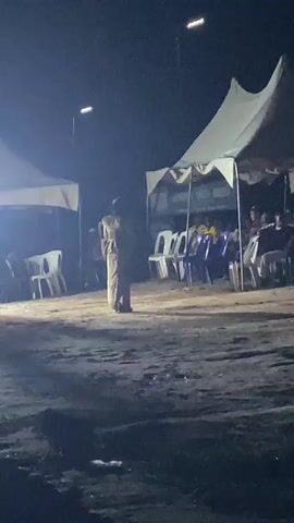 A dimly lit outdoor scene at night. In the foreground, a person stands facing away from the camera, wearing a light-colored garment. Behind this person, multiple white plastic chairs are arranged in rows under a large white tent. To the left, another tent with white fabric is visible, also with chairs underneath. The ground is unpaved and appears to be dirt or sand. In the background, the night sky is dark, with visible streetlights in the distance. The scene suggests an outdoor event or gathering taking place after dark, possibly a performance or a ceremony.