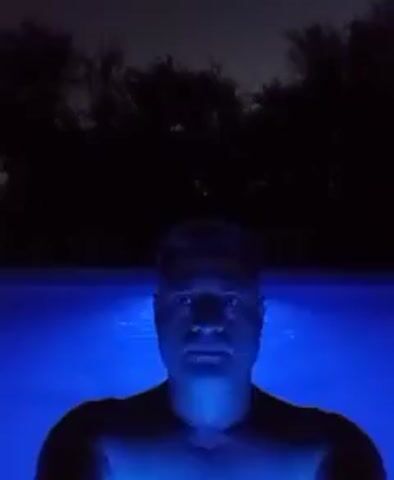 A man is standing in a pool lit with blue underwater lights at night. His head and shoulders are visible above the water. The area beyond the pool is dark, with some trees silhouetted against the sky. The water surface is slightly rippled. The man appears to be looking straight ahead at the camera.