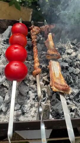 The video shows a close-up view of food being grilled over hot coals. The primary subjects are skewers of food on a grill. On the left, there are four bright red tomatoes on a skewer, glistening from the heat. Next to them, a skewer holds pieces of meat that appear to be chicken or lamb, twisted and browned from grilling. To the right, a skewer holds a portion of what looks like lamb ribs, with the bone exposed and the meat cooked to a golden-brown. Wisps of smoke rise from the grill, indicating the cooking process is underway. The background is slightly blurred but suggests an outdoor setting with greenery and possibly a structure, hinting at a garden or backyard barbecue. The lighting suggests it is daytime, likely a sunny day, given the bright colors and the quality of the light. The overall impression is one of a delicious outdoor meal being prepared. The location context provided is Toğanalı, Azerbaijan, which suggests a traditional Azerbaijani barbecue, known as a "kebab" or "shashlik." There is no visible text in the video.