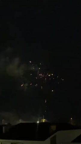 The video captures a nighttime scene with fireworks illuminating the dark sky. Several bursts of fireworks can be seen exploding and trailing smoke. In the foreground, the silhouettes of buildings with dark roofs are visible, with a few lights indicating structures and possibly streetlights. The fireworks display is the primary focus, with bright bursts of light and scattered sparks against the black backdrop of the night sky.