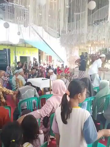 The video shows an indoor wedding ceremony. Guests are seated in rows of green plastic chairs, facing the front where a table with white cloth and microphones sits. Attendees are dressed in a variety of clothing styles, including traditional attire and modern casual wear. Some people are wearing head coverings. In the foreground, the backs of attendees’ heads are visible, including a girl with a ponytail. At the front, a speaker is addressing the gathering. Behind the speaker is a decorated stage area with flowers and white decorations. The ceiling is adorned with white streamers and round paper lanterns. The event is taking place under a temporary tent-like structure attached to a green-painted building. Some people are taking photos or videos with their phones.