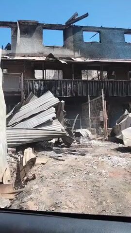 The video shows the aftermath of a fire. The building in the background is heavily damaged, with burnt walls and missing windows. Debris, including corrugated metal sheets, is scattered around the foreground. The footage is taken from inside a car, with a view of the devastated scene. A voice says, "There, our little house, our business, all lost."