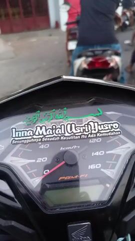 The video shows a first-person perspective from a motorcycle dashboard, looking forward at a line of motorcycles and riders on a paved street. The dashboard displays a speedometer with the needle indicating approximately 20 km/h. A sticker on the dashboard reads "Inna Ma'al 'Usri Yusro" in English and Arabic script, translating to "Verily, with hardship comes ease."

In the background, several people are on motorcycles, facing forward, suggesting they are in a queue or traffic. One rider in a black shirt is directly in front, their motorcycle's license plate reads "M 5086 AC." Other riders and motorcycles are visible further ahead and to the sides. They are on a street lined with buildings, one of which has red trim and appears to be a commercial establishment or office. The lighting suggests it is daytime, possibly late afternoon. The overall scene depicts a moment of stillness or slow movement in a line of traffic.