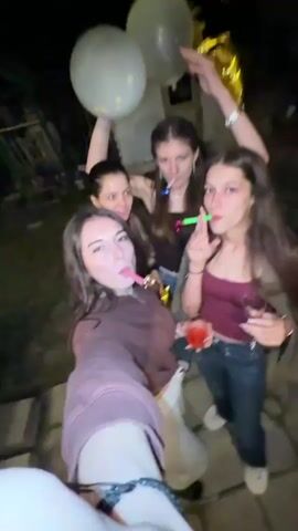 A group of five young women are celebrating outdoors at night. The woman closest to the camera is holding it in a selfie orientation, her arm outstretched, and blowing a pink party blower. To her right, a woman holds a red drink and a black party blower, also blowing it. Behind them, two women smile and hold party blowers, with one also holding a white balloon and the other holding a gold balloon. In the background, there is a dark, indistinct outdoor setting with some structures visible. The video is shaky and appears to be filmed with a handheld device.