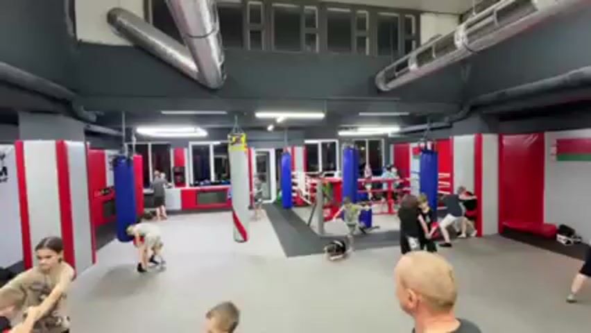 The video shows a wide shot inside a gym that appears to be for martial arts. There are a group of children in athletic clothing, some of whom are engaging in light sparring or wrestling on a mat. A man with a bald head and wearing a t-shirt stands in the foreground, observing the children. The gym has padded walls with red and white stripes, and several punching bags are hanging from the ceiling. There's a boxing ring in the background with people inside and around it. The room has a high ceiling with visible ventilation ducts. The lighting is bright and even throughout the space.