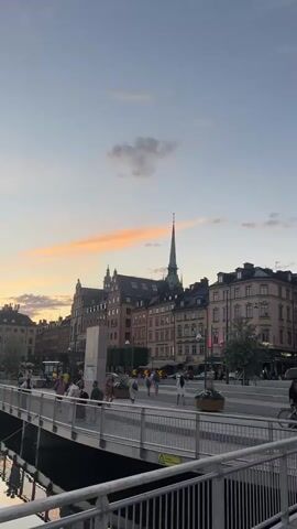 This is a vertical, short video of a scenic waterfront area in Stockholm, Sweden, likely during late evening or early morning hours. The sky displays a gentle gradient of blues, grays, and soft oranges, indicating twilight. A few wispy clouds are scattered across the sky, with a prominent band of orange clouds streaking horizontally near the horizon.

The foreground is dominated by a modern pedestrian bridge with a metal railing. People are casually walking across the bridge, enjoying the view. Below the bridge, a calm body of water reflects the sky and the buildings on the opposite bank. The water also shows reflections of several dark, rectangular barges or boats moored along the edge.

Across the water, a cluster of historic European-style buildings with pitched roofs lines the waterfront. One building features a tall, slender spire, characteristic of church architecture. Further down the waterfront, construction is visible, with scaffolding and cranes suggesting ongoing development or renovation. The overall atmosphere appears peaceful and serene, with the soft lighting and gentle movement of people creating a tranquil ambiance.

The video pans slightly, revealing more of the cityscape and the water. The time of day is clearly indicated by the soft, diffused light and the colors of the sky. The presence of water, historic architecture, and pedestrian activity strongly suggests an urban waterfront setting, consistent with Stockholm. There is no discernible text visible in the video.