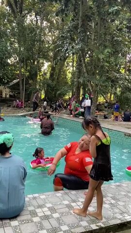 The video shows a crowded outdoor swimming pool area surrounded by lush green trees. In the foreground, a woman in an orange shirt and a young girl in a black swimsuit are near the edge of the pool. The girl is bent over, appearing to interact with something near the water. Another young girl in a pink swimsuit is floating on a watermelon-patterned inner tube in the pool. Several other people, including children and adults, are visible in and around the pool, some of whom are also using inflatable inner tubes. In the background, more people are gathered, with some sitting on the edge of the pool and others standing. The atmosphere appears to be a lively, recreational gathering.