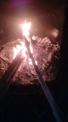 The video shows a close-up view of a fire burning inside what appears to be a dark, enclosed space, possibly a fire pit or stove. Flames flicker between two logs set in a teepee formation. Around the base of the fire are hot embers and ashes. The scene is dimly lit, emphasizing the bright light from the flames.