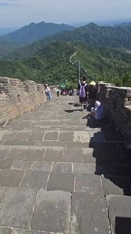 The video shows a descending view of a stone staircase on the Great Wall of China. The stone steps are large and rectangular, with brick walls on either side. Several tourists are visible on the steps and the walkway above. Some are walking down, while others are resting or looking at the scenery. The surrounding environment is mountainous and covered in lush green vegetation. The sky is clear and blue, indicating a sunny day. The camera perspective is that of someone walking down the stairs.