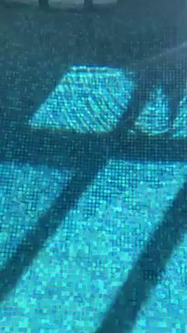 The video shows a small, indoor swimming pool with blue tile flooring. The water is clear and reflects light, creating patterns on the bottom of the pool. A large, multi-pane window looks out onto a grassy area with a few buildings and trees in the background. The window frame is made of dark wood. There are white lounge chairs visible on the ground outside the window. The ceiling is white with circular light fixtures.