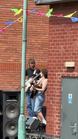 A man and a woman are performing music outdoors in front of a brick building. They are both playing guitars and appear to be singing. A large speaker is visible on the left side of the frame. Colorful bunting is strung across the top of the scene. The lighting suggests it is daytime.