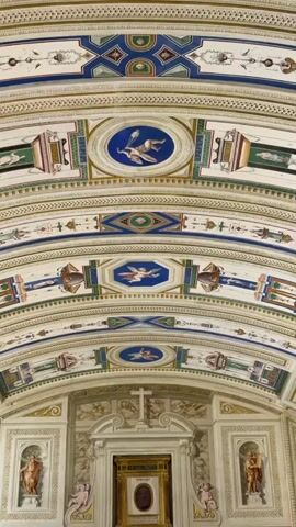 The video shows a close-up, upward view of an elaborately decorated ceiling. The ceiling is arched and features intricate patterns and paintings. The primary colors used are white, gold, blue, and green. There are multiple decorative bands running parallel to each other, adorned with geometric shapes, floral motifs, and small circular or octagonal medallions containing painted figures. These figures appear to be cherubs or putti in various poses, often depicted with wings. The overall impression is one of classical or Renaissance-inspired artistry. The lighting is even, suggesting an indoor setting with ample illumination. There are no people visible in the frame, and the focus remains solely on the architectural details of the ceiling.