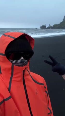 A person stands on a black sand beach wearing a bright orange hooded jacket, a black beanie, sunglasses, and a striped face covering. They are facing the camera. Another gloved hand appears from the side and flashes a peace sign. Behind the person are ocean waves, a dark, overcast sky, and rocky cliffs in the distance. The lighting is dim due to the cloudy weather.