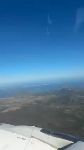 From an airplane window, a view of land with mountains and bodies of water under a clear, blue sky. A portion of the plane's wing is visible in the lower part of the frame with "NO STEP" printed on its surface. The terrain appears to be a mix of green fields and darker, possibly forested, areas. The water bodies are light blue and dotted with islands. The sky is cloudless.