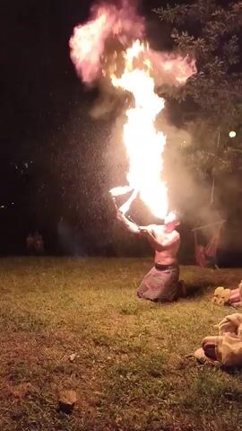 The video shows a person performing with fire at night in an outdoor grassy area. The person is wearing loose-fitting pants and is shirtless, with a lei or garland around their neck. They are holding two flaming objects, one in each hand, and appear to be juggling or spinning them. The fire illuminates the immediate surroundings, casting a warm glow on the person and the ground. In the background, there are trees and some faint lights, suggesting it is a park or an open area with some distant illumination. The overall atmosphere is that of a performance or a demonstration involving fire arts.