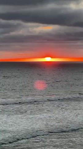 A slow-motion video shows a sunset over the ocean. The sun is a bright orange disk just above the horizon, casting a reflection on the water. Dark clouds are in the sky. Waves are breaking on the shore.