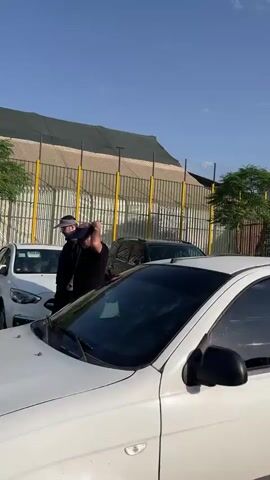 The video shows a street scene with several cars parked on the asphalt. A woman in a black hijab and abaya walks from right to left in the foreground, near a white car. Behind her, more white and silver cars are parked, with a fence and a building in the background. In the middle ground, a man with a red flag walks from left to right, followed by another person carrying a similar flag. Further back, a group of people, including a child in a wheelchair, are walking, also carrying red flags. The sky is blue with some clouds. The overall impression is of a procession or event taking place on a street.