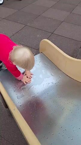 A toddler, dressed in a bright pink sweatshirt and grey pants, is on all fours at the bottom of a small, wavy plastic slide. The slide's surface is wet, with several puddles reflecting the overcast sky. The child's hands are spread out on the slide, and they appear to be pushing themselves up. Their hair is a light blonde.

The setting is a playground with a rubberized, patterned surface. In the background, a white and yellow tricycle with a black canopy is parked. Behind the tricycle, there are apartment buildings, suggesting an urban or suburban environment. A seesaw is visible in the distance, along with some greenery and trees. The overall lighting suggests it is daytime, but the cloudy sky indicates overcast weather. The child's focused expression suggests curiosity and engagement with the wet slide. There is no visible text in the media file. The location is identified as Myza (Inshinskii S/O), Russia.