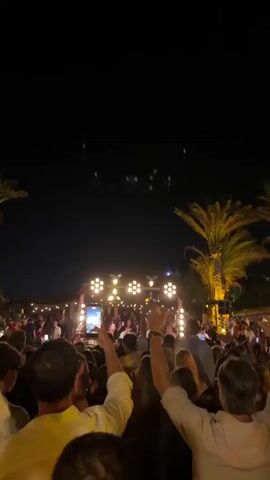 The video shows a crowded outdoor scene at night, likely a concert or festival. The sky is dark with a few small, greenish lights visible in the upper portion. In the midground, a stage or performance area is illuminated by bright lights. A large crowd of people is gathered in front of the stage, many of whom are holding up their phones to record. Palm trees with bright yellow lights illuminating them are visible on either side of the frame, suggesting a warm climate. The people in the foreground are mostly seen from behind, with their arms raised, some clapping. The overall atmosphere appears to be festive and energetic.