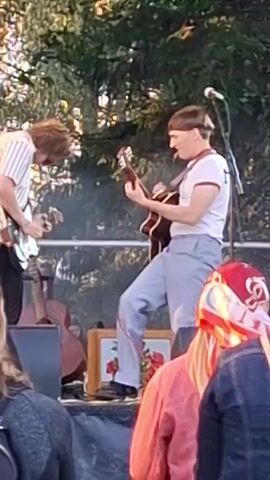 A live outdoor musical performance is captured from behind the audience. Two male musicians are on stage. The musician on the left wears a striped shirt and plays a guitar. The musician on the right wears a white t-shirt with red trim and light blue pants, and plays an acoustic guitar while seated on a stool. Both are positioned towards the center of the frame. Trees and foliage form the background. The foreground shows the backs of audience members, with some of their hair and shoulders visible. A microphone stand is visible on the right side of the stage. Red and white fabric is draped over an object in the lower right corner.
