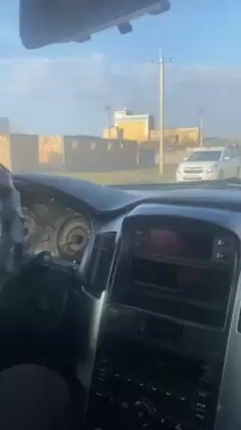 The view from inside a car shows a man walking in front of another white car. The road is narrow and residential, with buildings visible in the background. The sky is blue and partly cloudy. The car's dashboard is black and silver. A phone screen displaying an "abonent" (subscriber) message is visible.