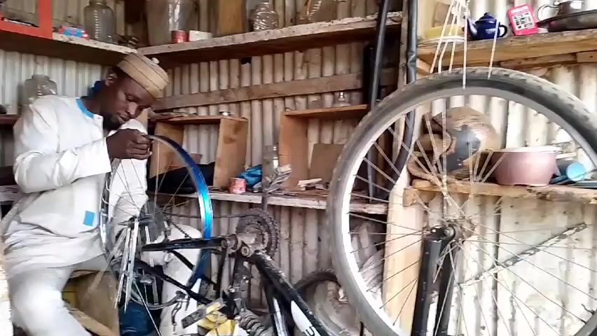 Here is a concise description of the video:

A man wearing a light-colored traditional outfit and a small, patterned cap is focused on repairing a bicycle wheel in a small, cluttered workshop. He is seated and working on a wheel mounted in a jig on a partially disassembled bicycle. Another bicycle wheel is hanging nearby. The workshop has corrugated metal walls and numerous shelves stocked with bottles, jars, tools, and other miscellaneous items.