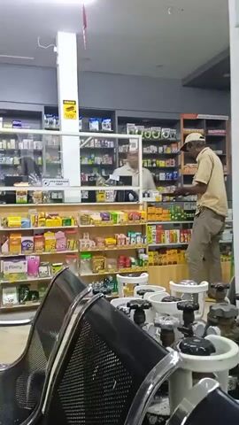 The video captures an indoor scene within what appears to be a pharmacy or convenience store in Kebonsari, Indonesia. In the foreground, several black metal mesh chairs with silver frames, typical of a waiting area, are visible. Immediately in front of these chairs, several small white objects with black tops and handles, resembling gas cylinders or industrial valves, are arranged on the floor.

Two men are the main subjects. On the right, a man with dark skin, wearing a light-brown collared shirt, light trousers, and a light-colored cap, stands in the aisle. He is meticulously counting what appears to be money in his hands. Behind him and slightly to the left, a man with glasses and a light-colored shirt is visible behind a service counter, which has a clear protective barrier. He appears to be working or observing.

The store is well-lit and features numerous shelves stocked with a wide variety of packaged products, including what look like food items, drinks, and possibly medicines, contributing to the retail environment. Visible text includes a "CCTV" sign on a pillar and "PENYERAHAN OBAT" (DRUG DISPENSING) on the counter, further suggesting a pharmaceutical context. The time of day is not explicitly shown but the interior is brightly lit by artificial light.
