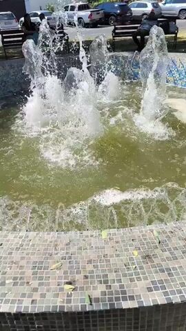 The video shows a fountain in an outdoor setting. The water in the fountain is murky green and splashes upwards in several jets. The edge of the fountain is tiled with small, multi-colored square tiles. In the background, there are people sitting on benches, trees, and several parked cars. The overall impression is of a public space on a sunny day.